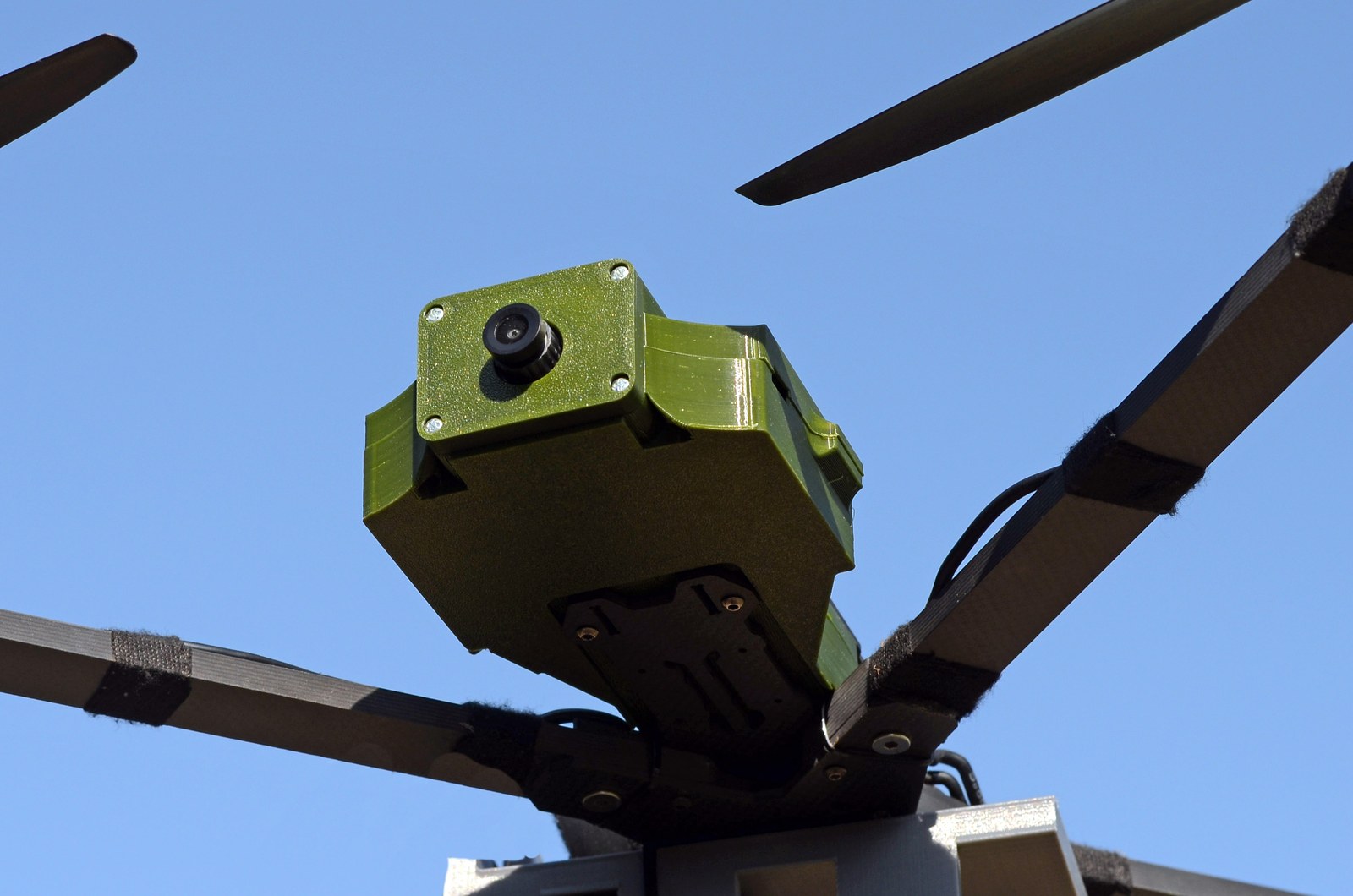 T-REX — FPV-рама в польоті, олів-зелений модуль донаведення з камерою, 3-лопатеві пропелери на тлі неба.